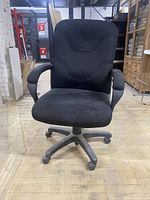 Front view of black office desk chair showing seat, backrest, armrests, and caster base in a warehouse setting.