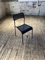 Single black plastic chair with perforated seat and backrest shown from side angle on wooden floor with brick wall background.