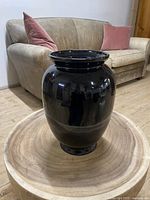 Full view of glossy black ceramic vase placed on wooden surface with couch in the background.