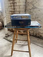 Three boxed board games stacked on a wooden chair in front of a white brick wall.