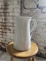 Full side view of the white ceramic pitcher on a wooden stool against a textured white brick wall background.
