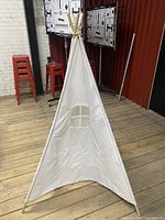 Full view of the children's white fabric teepee play tent with wooden poles on a wooden floor, showing its shape and window detail.