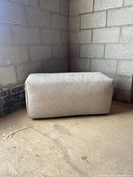 Side angle view of fabric ottoman showing texture and shape in a corner with concrete block walls.
