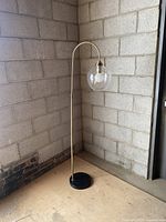 Full view of the lamp showing the curved gold-tone metal stand and black base with the clear glass globe shade that houses the bulb.