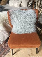 Grey fuzzy square pillow resting on an orange upholstered chair with black legs, shown from the front angle.