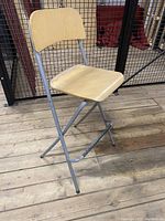 High stool with wooden seat and backrest, steel frame and legs, placed on wooden floor against metal mesh background.