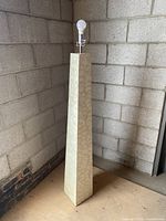 Tall rectangular tapered floor lamp with an ivory patterned base standing against a concrete block wall, with a light bulb installed and wire cut.