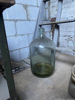 Photo of the large vintage green-tinted glass bottle sitting on the floor next to a metal table and a wall with cinder blocks and a step ladder.