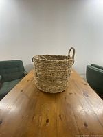 Wicker basket shown from front angle on wooden table with two chairs in background. Basket has one intact handle and one broken handle.