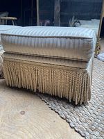 Side view of one foot stool showing striped cushion and tassel fringe base on wooden floor.