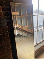 Photo of one full-length mirror leaning against a brick wall near a window, showing reflective surface and gold-tone frame.