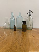 Photo of six vintage glass bottles on a wooden surface against a plain wall, showcasing variety in size and color.