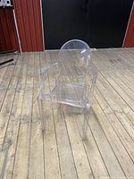 Side angled view of the transparent plastic chair showing its rounded backrest, armrests, and seating area.