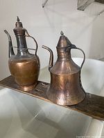 Two vintage copper kettles displayed side by side on a narrow wooden beam, showing overall condition and wear.