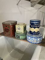 Front view of five assorted vintage metal tins placed on a shelf showing varying colors and designs.