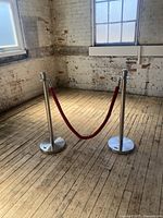 Two stainless steel stanchion posts with weighted round bases connected by a red velvet rope placed on wooden floor in front of white brick wall.