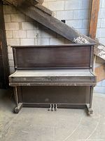 Front view of the vintage upright piano showing full face and pedals, wood finish dusty and worn in places