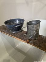 Metal sifter and colander shown on a wooden plank shelf, displaying overall shape and condition.