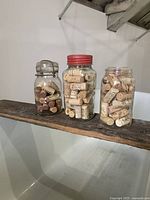 Photo shows three vintage glass jars filled with assorted wine corks, placed on wooden shelf against white wall.
