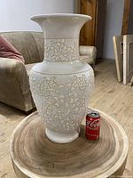 White ceramic vase with gold leaf pattern, shown on wooden surface with sofa in background and soda can for size reference.