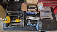 Two air nailers and assorted nails and staples laid out on and beside table, including carrying cases and open boxes of fasteners.