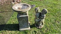 Wide view of bird bath on pedestal and cement statue of two children on grass outdoors.