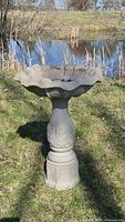 Full view of the cement bird bath standing on grass with pond and trees in background.