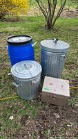 Photo showing three metal cans, a blue plastic barrel, and a brown lockbox on grass outdoors.