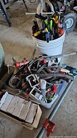 Various types and sizes of clamps piled in a plastic bin and a white bucket, including C-clamps and bar clamps visible at bottom and side.