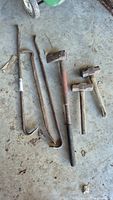 Full view of three crowbars, two mallets, and one axe arranged on a concrete floor showing rust and wear.