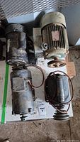 Four assorted electric motors placed on a surface with visible wiring, pulleys, and metal housings, showing signs of wear.