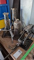 Photo showing two rusted metal jack stands, hydraulic bottle jack, and scissor jack on a wooden surface.