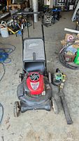 Photo showing red and black Craftsman Platinum lawnmower with collection bag and adjacent green Weedeater leaf blower on concrete floor indoors.