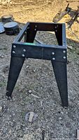 Black metal power tool base stand on dirt ground seen from side angle showing riveted construction and angled legs.