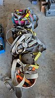 Buckets filled with multiple webbed straps in colors gray, yellow, orange, red, coiled and loose, alongside metal hooks and ratchets.