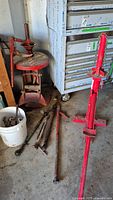 Two red manual tire changers with circular bases and lever arms, surrounded by multiple loose metal rods and arms on concrete floor near metal tool cabinet.