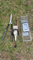 Three items laid out on grass: Fiskars clippers, IDC electric trimmer, and metal live capture trap