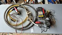 Full view of all items in the lot including the Biltek diesel fuel transfer pump with attached hoses and nozzle, battery tester, air pump with gauge, chain grinder, and soldering iron on a grey table.