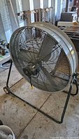 Front angled view of large industrial electric fan on black wheeled frame showing protective mesh grille, blades, and power cord.