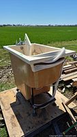 Full view of walk-in tub showing the basin, faucet, hose, and outer beige body. Positioned outdoors on wooden planks with green field background.