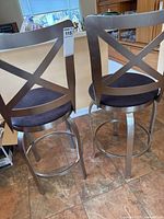 Pair of metal frame barstools with X-back design and fabric covered seats seen from the back.
