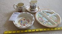 Photo showing two Bunnykins mugs, a Bunnykins bowl, and a divided Bunnykins plate on carpet with measuring tape for scale