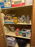 Shelf with multiple boxes and loose Mason jars, lids, a metal strainer, and a black canning pot at bottom.