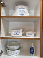 Three shelves inside a cabinet showing two white Pyrex casserole dishes with blue cornflower design on top shelf, two white CorningWare dishes with floral pattern and some lids on middle shelf, and one smaller white CorningWare dish with lid plus Pyrex cleaner bottle on bottom shelf.