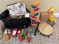KNEX building parts and instruction booklet in a black bin, small toy cars arranged in front, wooden/plastic marble run, tambourine, hand drum, yellow doll with hood.