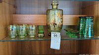 Full display showing large decorated vase, six small floral glasses, green decorative bottle, and three green glass cups with saucers on a glass shelf.