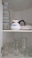 View of white Corning Ware teapot on shelf with glass pitchers, bowls, and small glass cups arranged below. Several stackable glass storage containers visible on left.