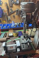 Front and top view of GH 1/2 HP drill press and Black & Decker bench grinder side by side on workbench, showing drill chuck, vise, and grinder discs.