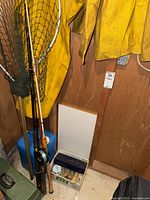 Photo showing yellow waterproof jacket and pants hanging on the wall, fishing rods leaning against the wall, fishing net, and white tackle box with fishing accessories on the floor.