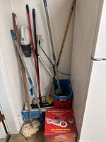 Photo of cleaning items including Ultimate Spin Mop box, handheld vacuum, brooms, mop, bucket, and other cleaning tools in laundry room corner